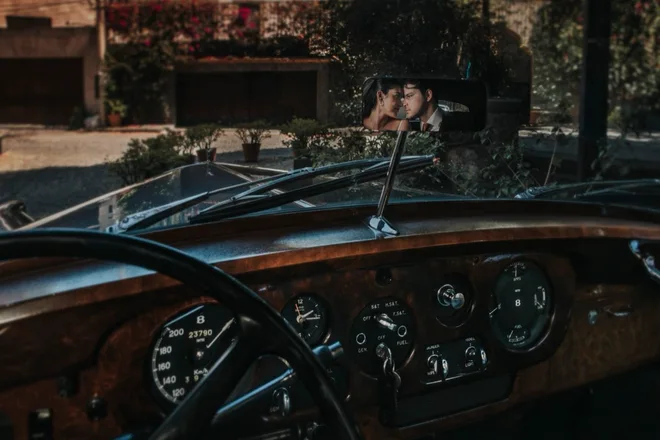 Reflejo de los novios Alex y Andy en el espejo de un auto clásico el día de su boda