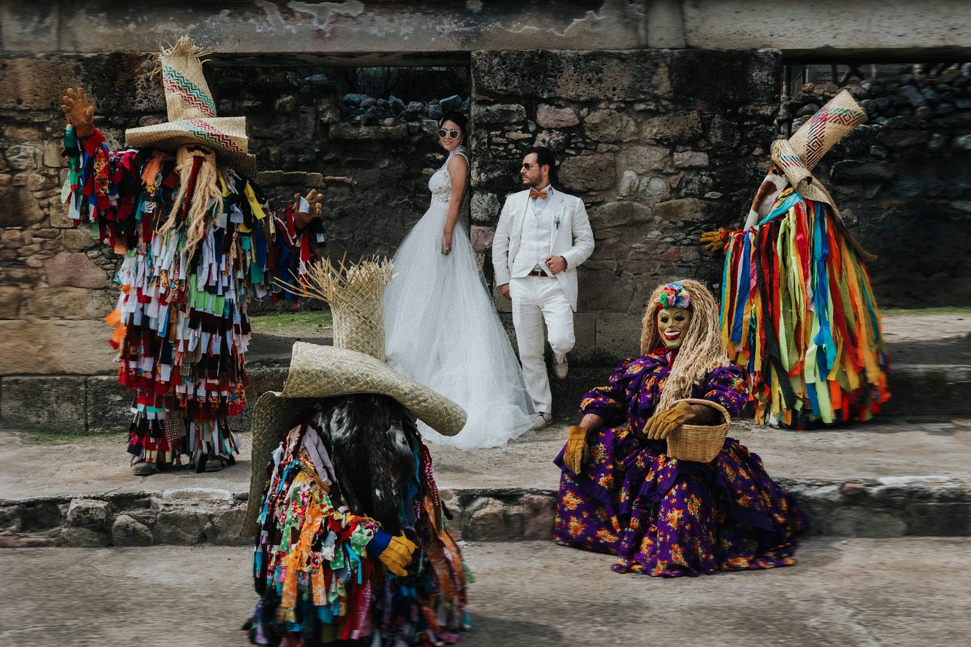Novios acompañados de tiliches, con sus trajes de tiras de tela y máscaras, junto a un muro de piedra zapoteca en Mitla, Oaxaca