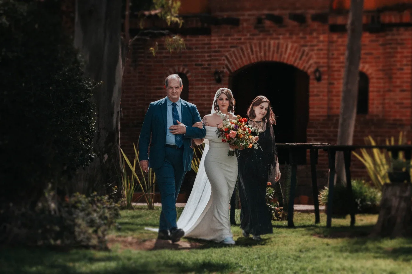 Lucía caminando hacia el altar acompañada de sus padres, con la arquitectura de ladrillo de Quinta San Carlos de fondo