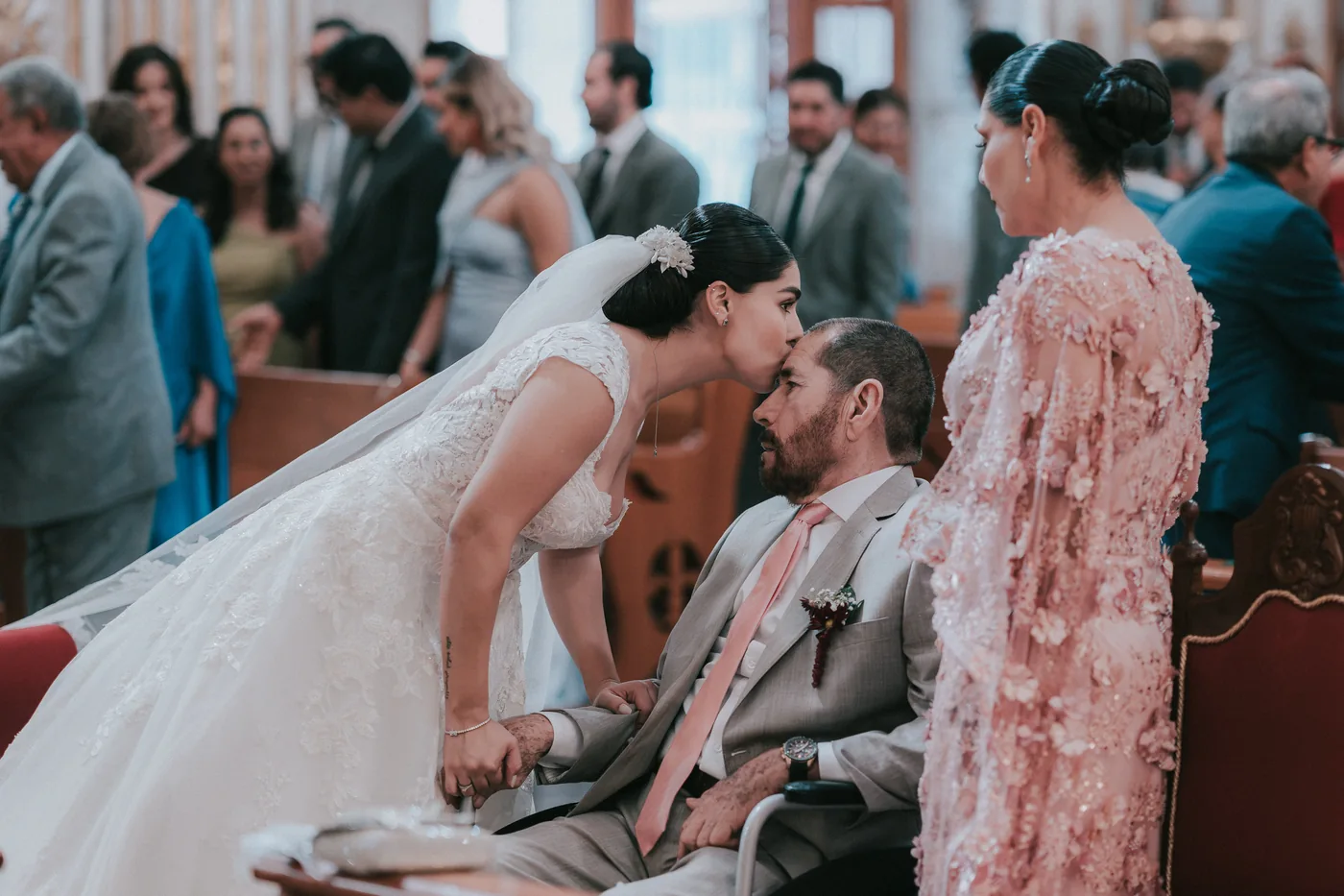 Marla se agacha a besar en la frente a su papá, que la espera en silla de ruedas junto al altar, con su mamá al lado