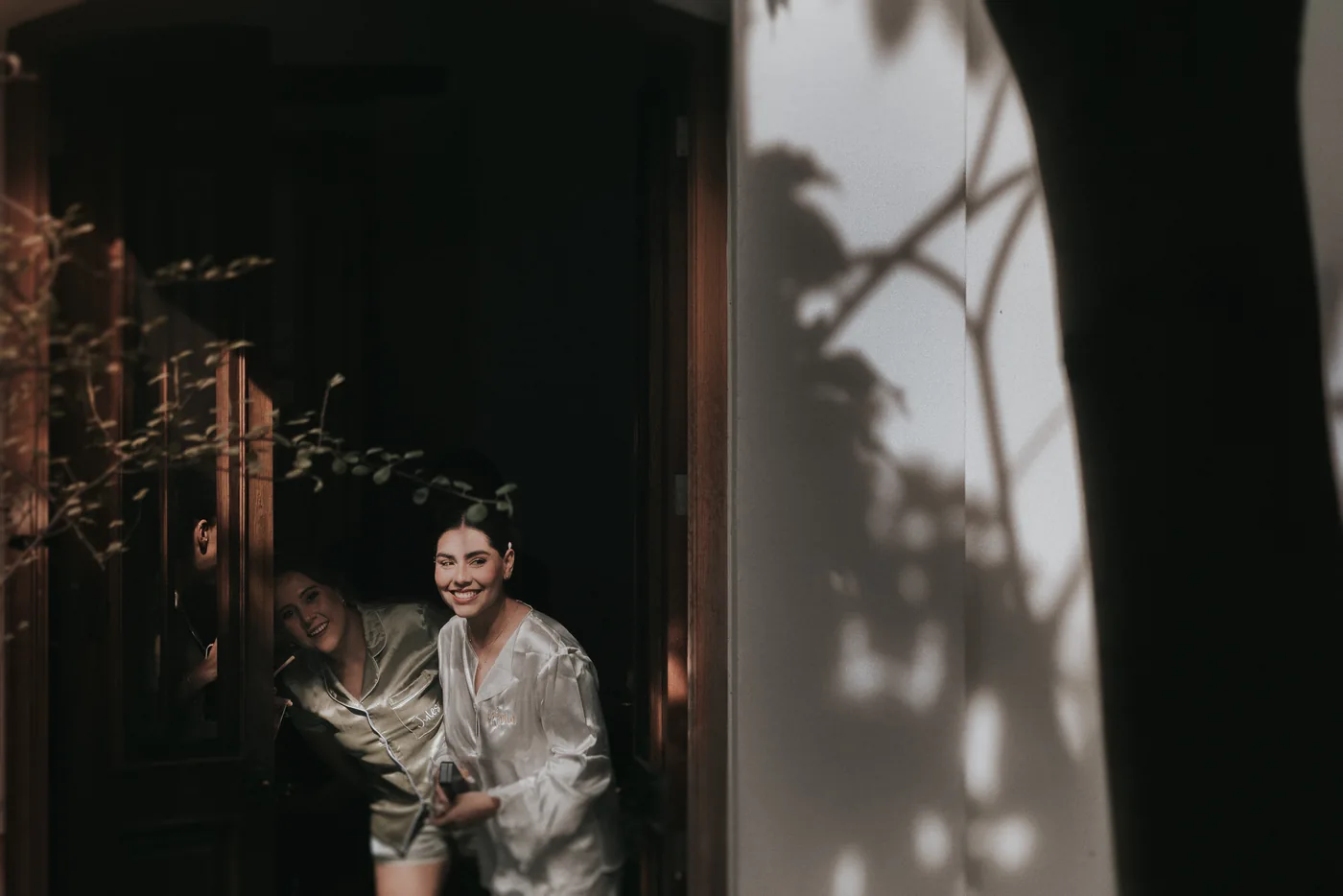 Marla, en pijama de satín blanco, se asoma sonriendo por una puerta de madera junto a una amiga