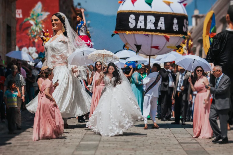 La novia caminando por las calles de Oaxaca durante la calenda, entre sombrillas y mojigangas