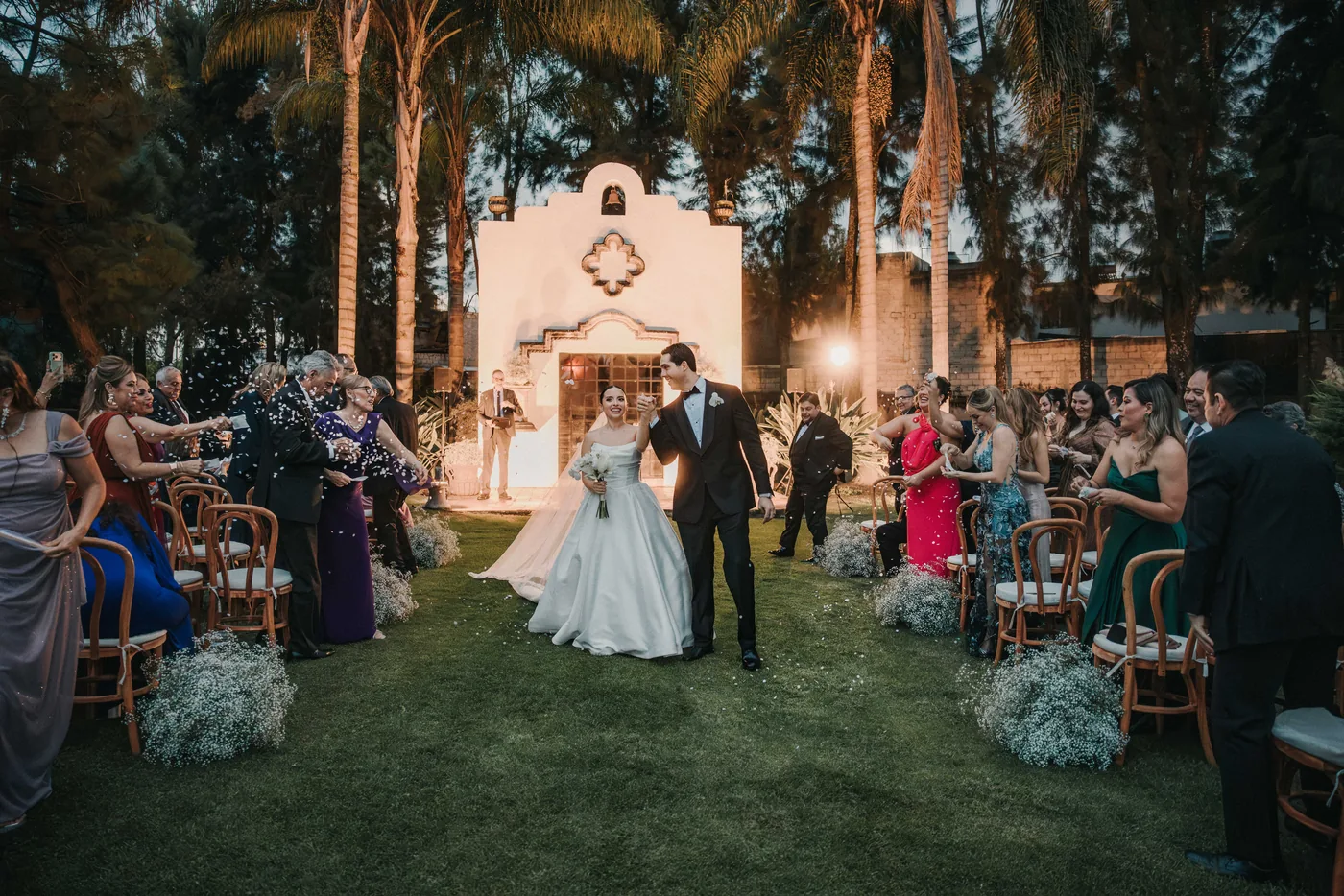 Sofi y Diego salen de la ceremonia entre los invitados, de noche, con la fachada blanca iluminada