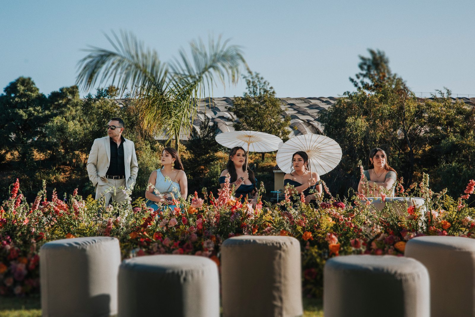 Invitados con sombrillas de papel esperando la ceremonia, detrás de un muro de flores